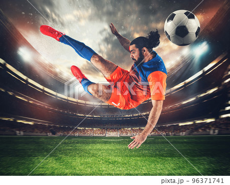 Soccer striker with orange and blue uniform hits the ball with an acrobatic kick in the air at the stadium 96371741