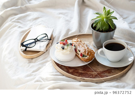 Cakes, coffee cup, notebook and eyeglass on wooden tray on bedding. Cozy breakfast in bed. 96372752