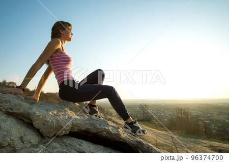 Woman hiker sitting on a steep big rock enjoying warm summer day. Young female climber resting during sports activity in nature. Active recreation in nature concept. 96374700