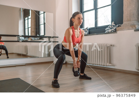 A strong, muscular female weightlifter is doing squats with a kettlebell in a gym. A strong, muscular female weightlifter is doing squats with a kettlebell in a gym. 96375849