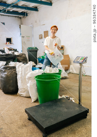 Vertical portrait of smiling female worker in latex gloves looking at camera sorting diverse waste for further disposal at private waste recycling plant. Concept of recycling and garbage sorting Vertical portrait of smiling female worker in latex gloves looking at camera sorting diverse waste for further disposal at private waste recycling plant. Concept of recycling and garbage sorting 96384740