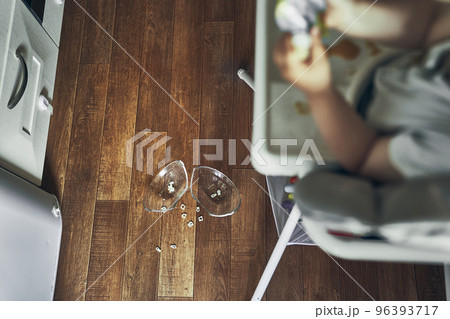 A small baby on a high chair dropped a cup of noodle soup on the wooden floor. The transparent plate is divided into two parts. Noodles in the form of figures. The table is dirty. High quality photo 96393717