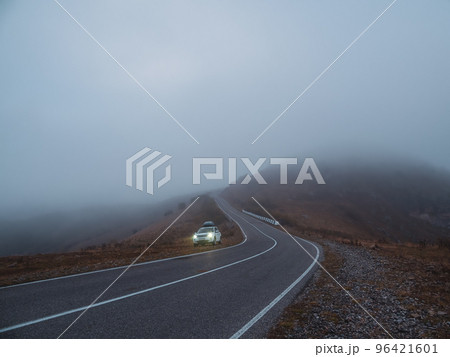A Rover car SUV with fog lights turned on is parked next to a scenic route road in a foggy autumn landscape. Luggage trunk box mountain on car roof rack. A Rover car SUV with fog lights turned on is parked next to a scenic route road in a foggy autumn landscape. Luggage trunk box mountain on car roof rack. 96421601