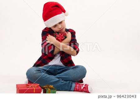 Adorable child boy in Santa hat, gently hugging a Christmas gift, isolated over white background, copy advertising space. Boxing Day. Happy festive atmosphere of winter holidays. New Year celebration 96423043