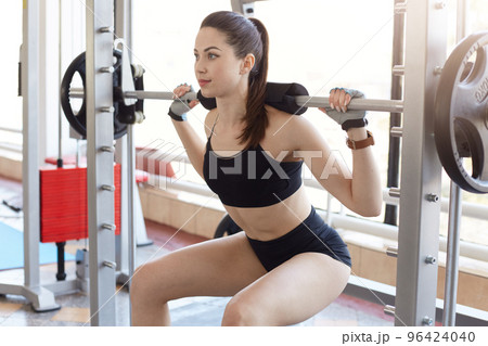 Athletic strong young lady doing back squat, spending her free time in gym, keeping her body in perfect form, wearing black tight sports suit. People, healthy lifestyle and sport concept. Copy space. Athletic strong young lady doing back squat, spending her free time in gym, keeping her body in perfect form, wearing black tight sports suit. People, healthy lifestyle and sport concept. Copy space. 96424040