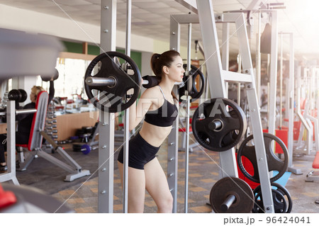 Young woman in gym doing squats with extra weight on shoulders, beautiful female working out at health club, woman develop definition of muscles developing definition of muscles. 96424041