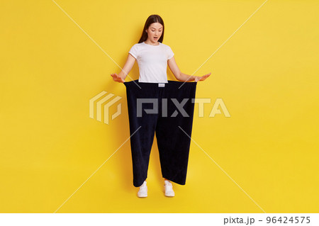 Full length photo of attractive young female shows her old huge pair of pants, looks shocked and surprised, looking down with opened mouth, lady after weight loss posing isolated on yellow background 96424575