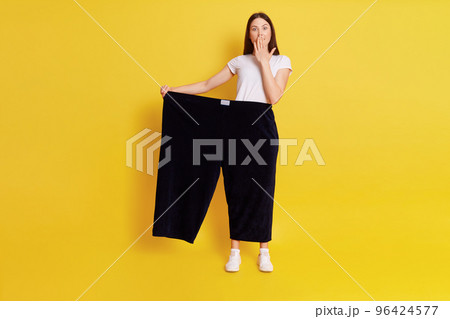 Full length of Caucasian astonished female lost weight and wearing old pants, being shocked and covering mouth with palm, posing isolated over yellow background. 96424577
