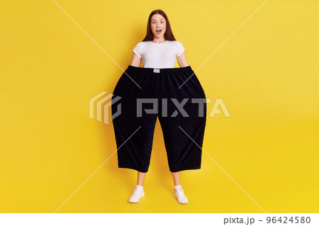 Shocked astonished woman wearing old too big black trousers keeps hands in pants. looks at camera with open mouth and big eyes, has surprised facial expression, posing isolated over yellow background. Shocked astonished woman wearing old too big black trousers keeps hands in pants. looks at camera with open mouth and big eyes, has surprised facial expression, posing isolated over yellow background. 96424580