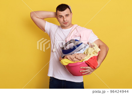 Indoor shot of puzzled handsome man busy with housework, does home chores, holds basin of laundry, isolated over yellow background. Tired male housekeeper keeps hand on head, being exhausted. Indoor shot of puzzled handsome man busy with housework, does home chores, holds basin of laundry, isolated over yellow background. Tired male housekeeper keeps hand on head, being exhausted. 96424944