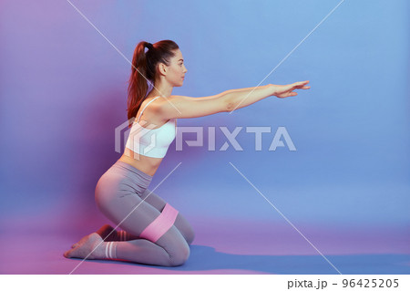 Side view of strong and fit athletic young caucasian woman in sportswear with bands training legs and glutes muscular while sitting on floor on knees, keeping hands raised. 96425205