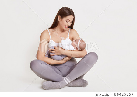 Mother holding newborn baby girl while sitting on floor with crossed legs, wearing white t shirt and gray leggins, posing against light background, smiling lady look at kid. Mother holding newborn baby girl while sitting on floor with crossed legs, wearing white t shirt and gray leggins, posing against light background, smiling lady look at kid. 96425357