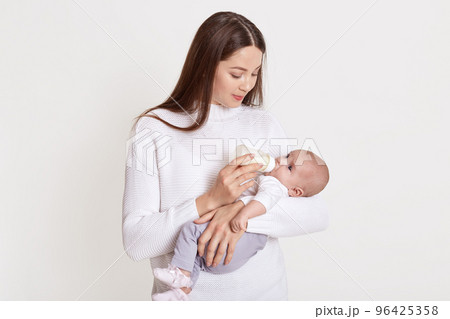 Mother feeding her baby from feeding bottle, dark haired female wearing sweater hold baby in hands and looking at her kid, posing isolated over white background. Mother feeding her baby from feeding bottle, dark haired female wearing sweater hold baby in hands and looking at her kid, posing isolated over white background. 96425358