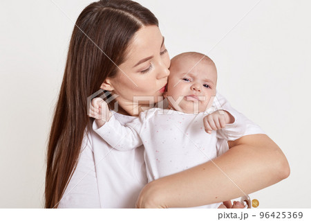 Mother kissing and hugging newborn daughter isolated over white background, tender, care, lady with brown hair expressing love to her child. 96425369