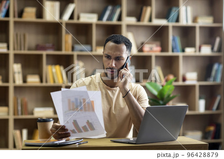 Irritated arab businessman working from home, sitting at desk with laptop, holding documents and having phone call Irritated arab businessman working from home, sitting at desk with laptop, holding documents and having phone call 96428879