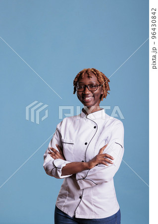 African american professional female chef posing in white uniform in studio shot. Happy cook with curly hair looking at camera with arms crossed against blue background. African american professional female chef posing in white uniform in studio shot. Happy cook with curly hair looking at camera with arms crossed against blue background. 96430842