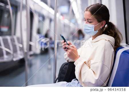 Active girl in a protective mask riding on a subway train with a mobile phone 96434358