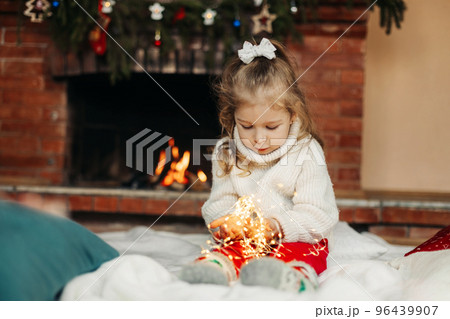 Cute little girl sitting by the fireplace with a Christmas garland.Christmas portrait, cozy style. The concept of Christmas and New Year 96439907