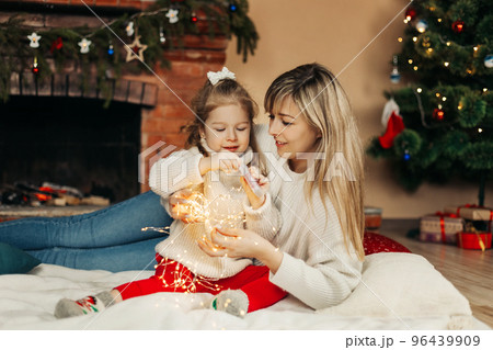 Cheerful mom and daughter play with Christmas lights on the floor by the fireplace, decorate the room for the holidays. The concept of Christmas and New Year 96439909