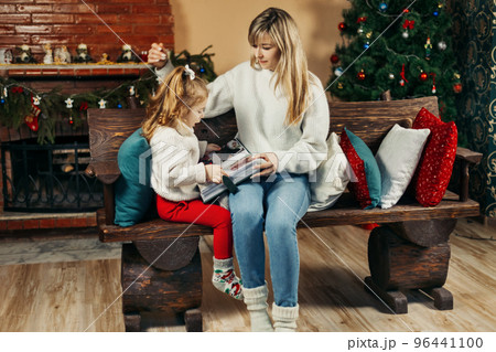 Cute mom and daughter are looking through a family photo album on Christmas Eve, spending time together by the fireplace and the Christmas tree. Christmas, New Year, memories, family evening 96441100