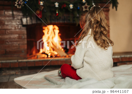 A happy little girl is sitting by the fireplace in a cozy living room on Christmas Eve. The concept of Christmas and New Year. Rear view A happy little girl is sitting by the fireplace in a cozy living room on Christmas Eve. The concept of Christmas and New Year. Rear view 96441107