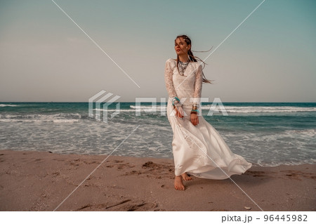 Model in boho style in a white long dress and silver jewelry on the beach. Her hair is braided, and there are many bracelets on her arms. 96445982