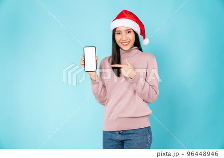 woman wearing red Christmas hat and holding smartphone and smiling on light blue background. woman wearing red Christmas hat and holding smartphone and smiling on light blue background. 96448946