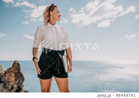 Digital nomad, Business woman working on laptop by the sea. Pretty lady typing on computer by the sea at sunset, makes a business transaction online from a distance. Freelance, remote work on vacation 96449791