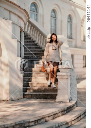 Woman walks in the city, lifestyle. Young beautiful woman in a loose light sweater, brown skirt and sneakers with a hat. Woman walks in the city, lifestyle. Young beautiful woman in a loose light sweater, brown skirt and sneakers with a hat. 96451144
