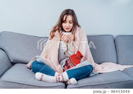 Woman freezes in wintertime. Young girl wearing warm woolen socks and wrapped into two blankets, holding a cup of hot drink and heating pad while sitting on sofa at home. Keep warm. Selective focus. 96453049