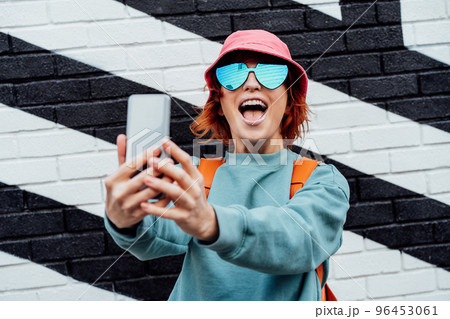 Excited redhead woman screaming while taking a selfie photo outdoors. Emotional hipster fashion women in bright clothes, heart shaped glasses, bucket hat taking selfie photo on the phone camera Excited redhead woman screaming while taking a selfie photo outdoors. Emotional hipster fashion women in bright clothes, heart shaped glasses, bucket hat taking selfie photo on the phone camera 96453061