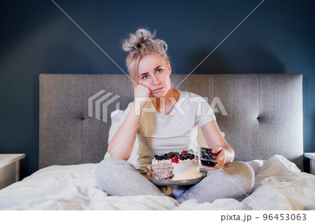 Bored, sad woman in home clothes eating cake and using the remote control to change the channel while watching television in bed at night. Overeating, Jamming of negative emotions. Selective focus. Bored, sad woman in home clothes eating cake and using the remote control to change the channel while watching television in bed at night. Overeating, Jamming of negative emotions. Selective focus. 96453063
