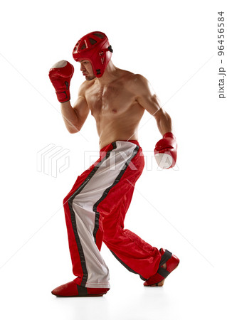 Dynamic portrait of young muscled athlete, professional kickboxer in protective helmet and boxing gloves in action isolated on white background. Sport, competition, energy, combat sports 96456584