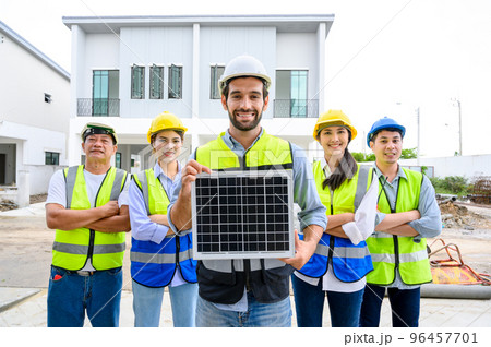 electrician man in safety vest with helmet showing solar cell panel with the team in background, standing in the under-construction building. Green energy electricity concept. concept of sustainable electrician man in safety vest with helmet showing solar cell panel with the team in background, standing in the under-construction building. Green energy electricity concept. concept of sustainable 96457701
