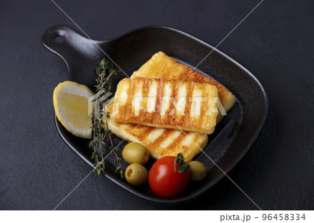 Grilled halloumi cheese in a black pan with olives, tomatoes, cucumbers, thyme and lemon. Traditional cheese of the Eastern Mediterranean. Black background, selective focus, close-up. Grilled halloumi cheese in a black pan with olives, tomatoes, cucumbers, thyme and lemon. Traditional cheese of the Eastern Mediterranean. Black background, selective focus, close-up. 96458334