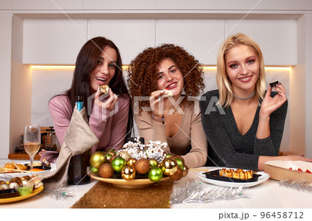 Three young cheerful intercultural women with alcoholic drinks standing in front of camera while enjoying home party in living room 96458712