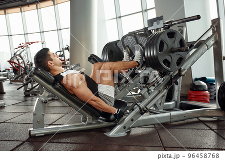 Man Using A Press Machine In A Fitness Club. Strong man doing an exercise on its feet in the simulator. Side view. 96458768