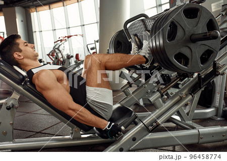 Young man is engaged on a modern simulator in the gym. Male athlete is doing exercises for legs with training machine. 96458774