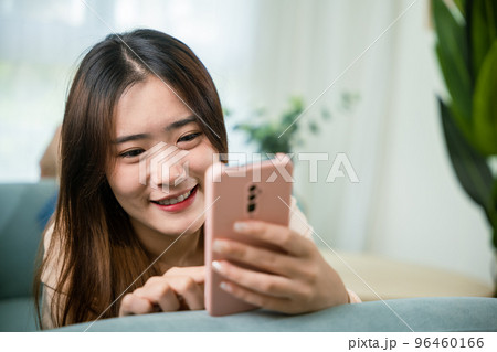 Smiling attractive woman relaxed reading text message on smartphone, Asian young female using smart mobile phone lying on sofa in living room room at home, gadget online application making internet Smiling attractive woman relaxed reading text message on smartphone, Asian young female using smart mobile phone lying on sofa in living room room at home, gadget online application making internet 96460166