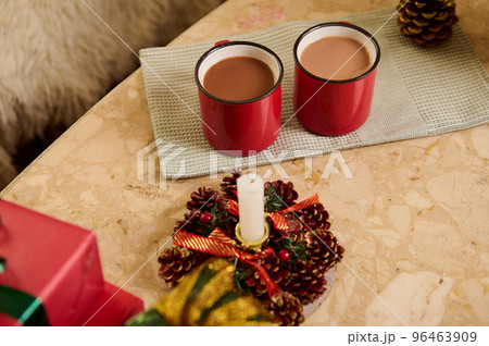 Close-up of two red cups with hot cocoa or chocolate drink on a green napkin, next to a decorative Christmas lit candle, pine cones and beautiful presents on marble table. New Year atmosphere at home. 96463909