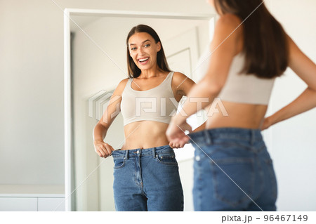Successful slimming concept. Excited caucasian lady wearing jeans after weight loss, comparing size, posing near mirror 96467149