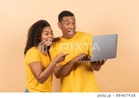 Portrait of excited black couple pointing at laptop together and looking at gadget with excitement, peach background Portrait of excited black couple pointing at laptop together and looking at gadget with excitement, peach background 96467438