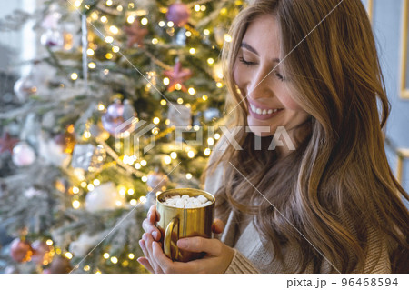 Closeup happy woman holding golden luxury mug with hot marshmallow cocoa, coffee at Christmas tree Closeup happy woman holding golden luxury mug with hot marshmallow cocoa, coffee at Christmas tree 96468594