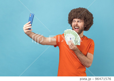 Portrait of excited crazy man with Afro hairstyle wearing orange T-shirt having video call or livestream, showing euro banknotes to subscribers. Indoor studio shot isolated on blue background. Portrait of excited crazy man with Afro hairstyle wearing orange T-shirt having video call or livestream, showing euro banknotes to subscribers. Indoor studio shot isolated on blue background. 96471576