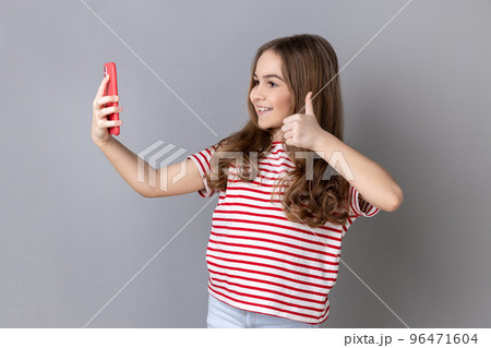Portrait of little girl wearing striped T-shirt making video call using smartphone, smiling with toothy smile, showing thumb up. Indoor studio shot isolated on gray background. 96471604