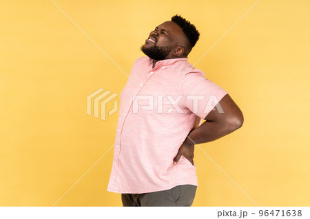 Back, kidney or spine pain. Portrait of man wearing pink shirt standing and endure pain on back, frowning face, suffering terrible pain. Indoor studio shot isolated on yellow background. Back, kidney or spine pain. Portrait of man wearing pink shirt standing and endure pain on back, frowning face, suffering terrible pain. Indoor studio shot isolated on yellow background. 96471638