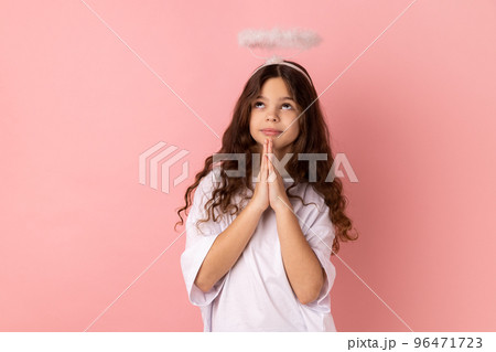 Portrait of little girl wearing white T-shirt keeps palms pressed together, pleading angel, has innocent expressions, asks for mercy or apologize. Indoor studio shot isolated on pink background. 96471723