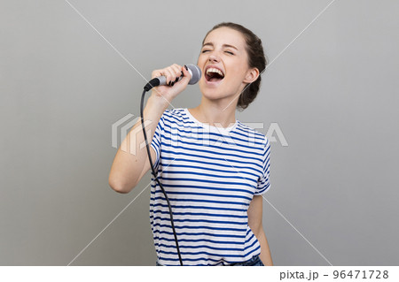 Portrait of excited positive woman wearing striped T-shirt singing songs at karaoke, having fun, holding microphone, entertainment. Indoor studio shot isolated on gray background. Portrait of excited positive woman wearing striped T-shirt singing songs at karaoke, having fun, holding microphone, entertainment. Indoor studio shot isolated on gray background. 96471728