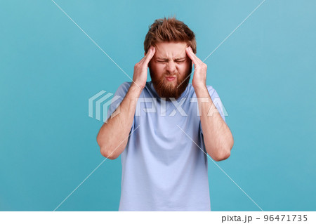 Portrait of unhealthy bearded man massaging temples feeling headache, suffering migraine or high blood pressure, trying to concentrate. Indoor studio shot isolated on blue background. 96471735