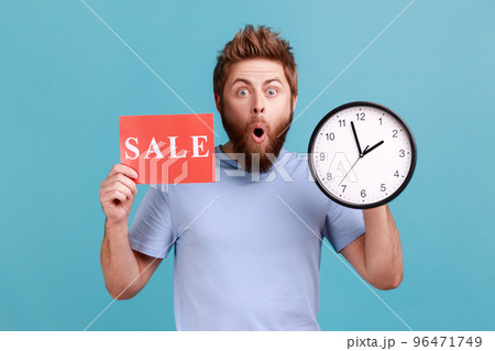 Portrait of shocked astonished handsome bearded man holding clock and card with sale inscription, announcing finishing huge discounts. Indoor studio shot isolated on blue background. 96471749